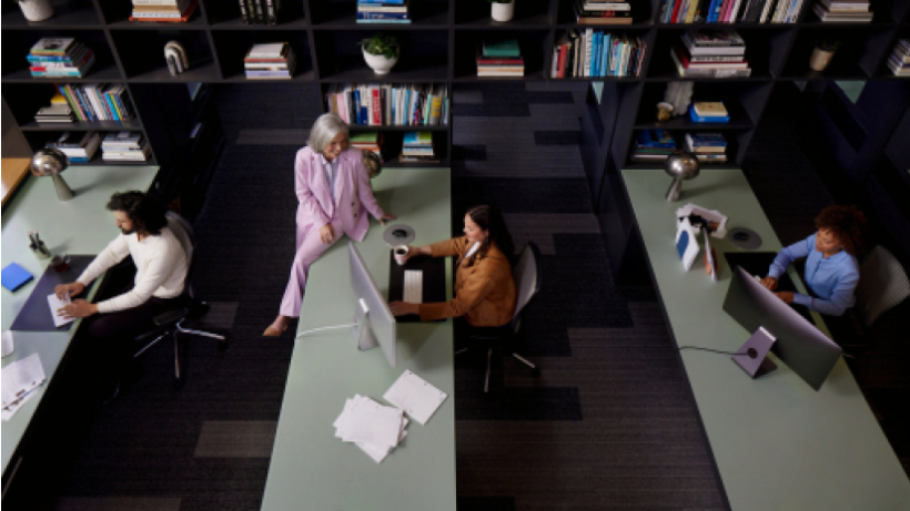 A woman chatting with someone working at a desk in a large open-plan office.